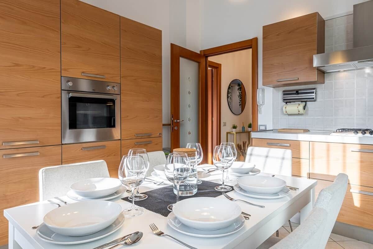 Een moderne keuken met houten kasten, een inbouwoven en een eethoek voor vier personen, compleet met witte borden en wijnglazen.