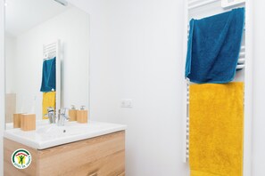 A bathroom with a white sink, wooden vanity, and a mirror. There are two towels, one blue and one yellow, hanging on a rack. - Les Dahus Tranquilles, All-Inclusive Vacation Rental (LE HAUT-BREDA)