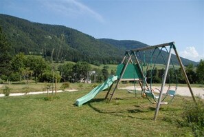 Zona de jocs amb tobogan i cadira de balanceig en un espai verd amb muntanyes al fons.