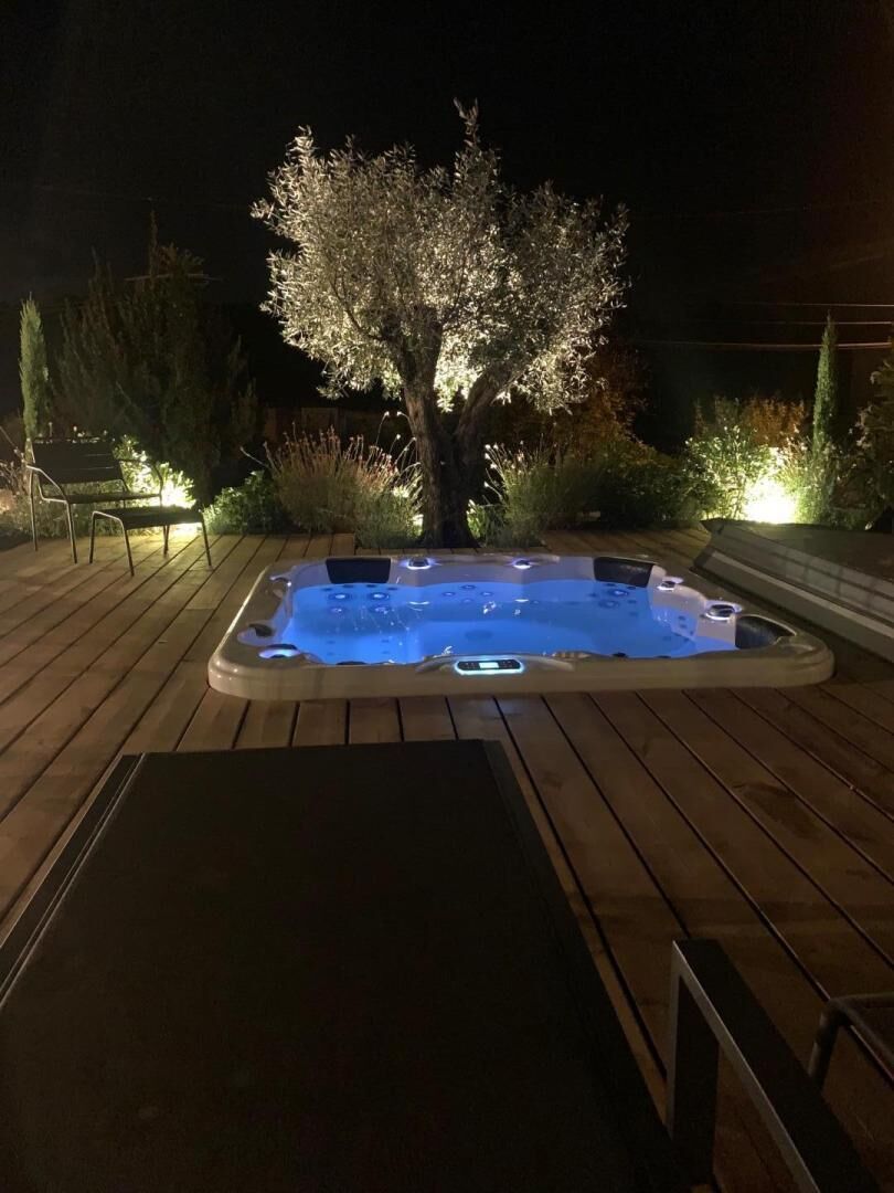 Ein Whirlpool auf einer Holzterrasse bei Nacht, umgeben von Pflanzen und einer Bank.