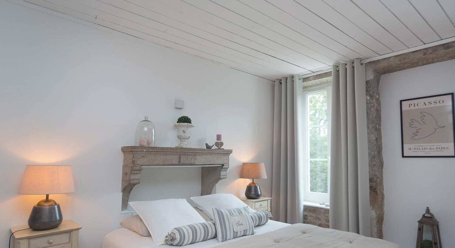 Ein Schlafzimmer mit einem Bett, zwei Nachttischen, einem Kamin, einem Fenster mit Vorhängen und einem gerahmten Kunstwerk an der Wand.