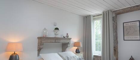 A bedroom with a bed, two bedside tables, a nightstand, a fireplace, a window with curtains, and a framed artwork on the wall.