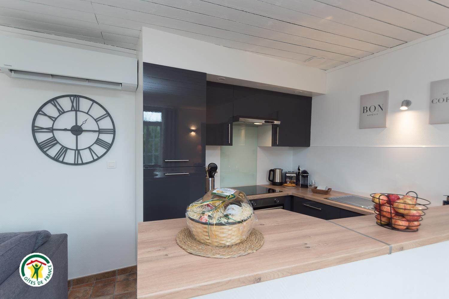A modern kitchen with a wooden countertop, a large clock, and a basket of fruits.