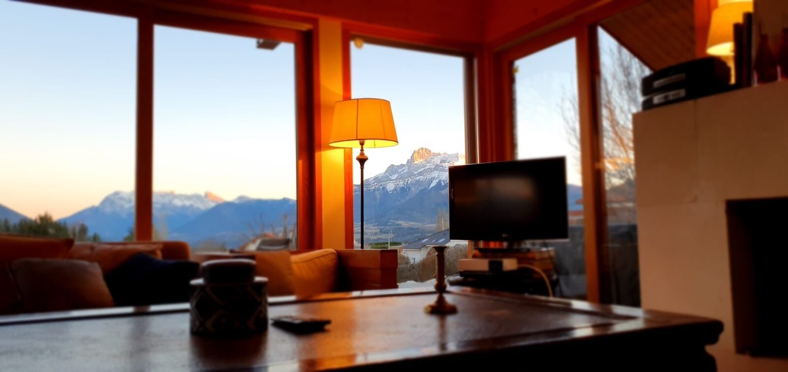 Una habitación con televisión, una mesa con lámpara y vistas a montañas a través de amplias ventanas.