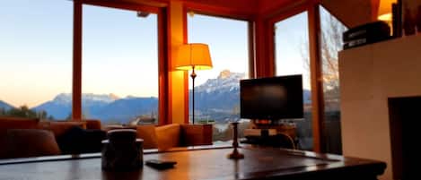 A room with a television, a table with a lamp, and a view of mountains through large windows.