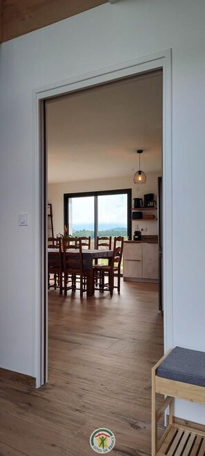 Ein Blick ins Zimmerinnere zeigt einen Holzboden, einen Esstisch mit Stühlen und einen Durchgang durch eine Glastür zu einem Küchenbereich mit einer Pendelleuchte.