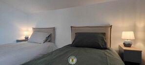 Ein Hotelzimmer mit zwei Betten, jeweils mit Kopfteil, Nachttisch und Lampe, sowie einem Logo auf der Bettdecke.