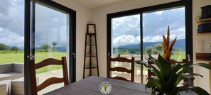 A room with a table, chairs, a plant, a shelf with a picture, and a view of a green landscape through large windows. - Mont Charvet - VELANNE (VELANNE)