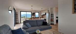 A modern living room with a grey sofa, a dining area with wooden chairs, and a kitchen with white cabinets. - Mont Charvet - VELANNE (VELANNE)