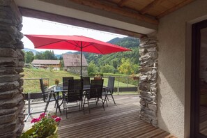 Ein überdachter Außenbereich mit Tisch und Stühlen, einem roten Sonnenschirm und Blick auf eine gebirgige Landschaft.