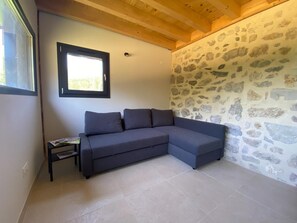 A room with a grey sectional sofa, a small table with magazines, a window with a black frame, and a stone-patterned wall. - The Three Som - SAINT PIERRE DE CHARTREUSE (SAINT PIERRE DE CHARTREUSE)