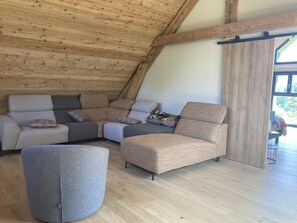 A loft-style room with a wooden ceiling and a large window. There is a sectional sofa with multiple cushions and a round ottoman. - The Three Som - SAINT PIERRE DE CHARTREUSE (SAINT PIERRE DE CHARTREUSE)