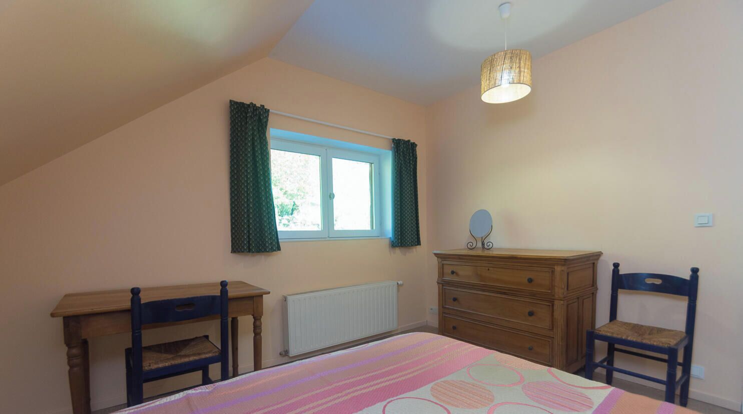A bedroom with a bed, a dresser, two chairs, a window with curtains, and a hanging light fixture.