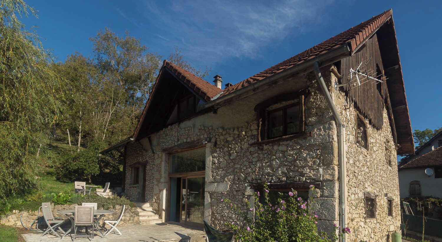 A stone building with a sloped roof, a door, and a window. There is a patio with chairs and a table.