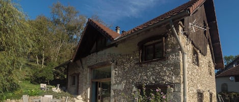 Un edificio de piedra con techo inclinado, una puerta y una ventana. Hay un patio con sillas y una mesa.