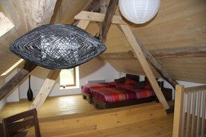 Une chambre à coucher aménagée dans un grenier, avec un plafond en pente, un lit recouvert d’une couvre-lit à rayures rouges et noires, et un luminaire suspendu en fibres tressées.