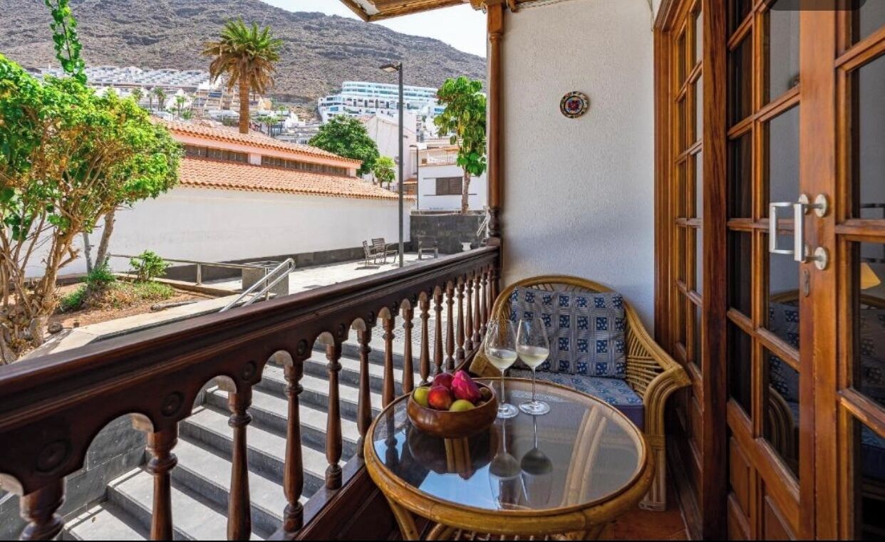 A balcony with a view of a cityscape, a round table with a bowl of fruit and two glasses, and a wooden door.