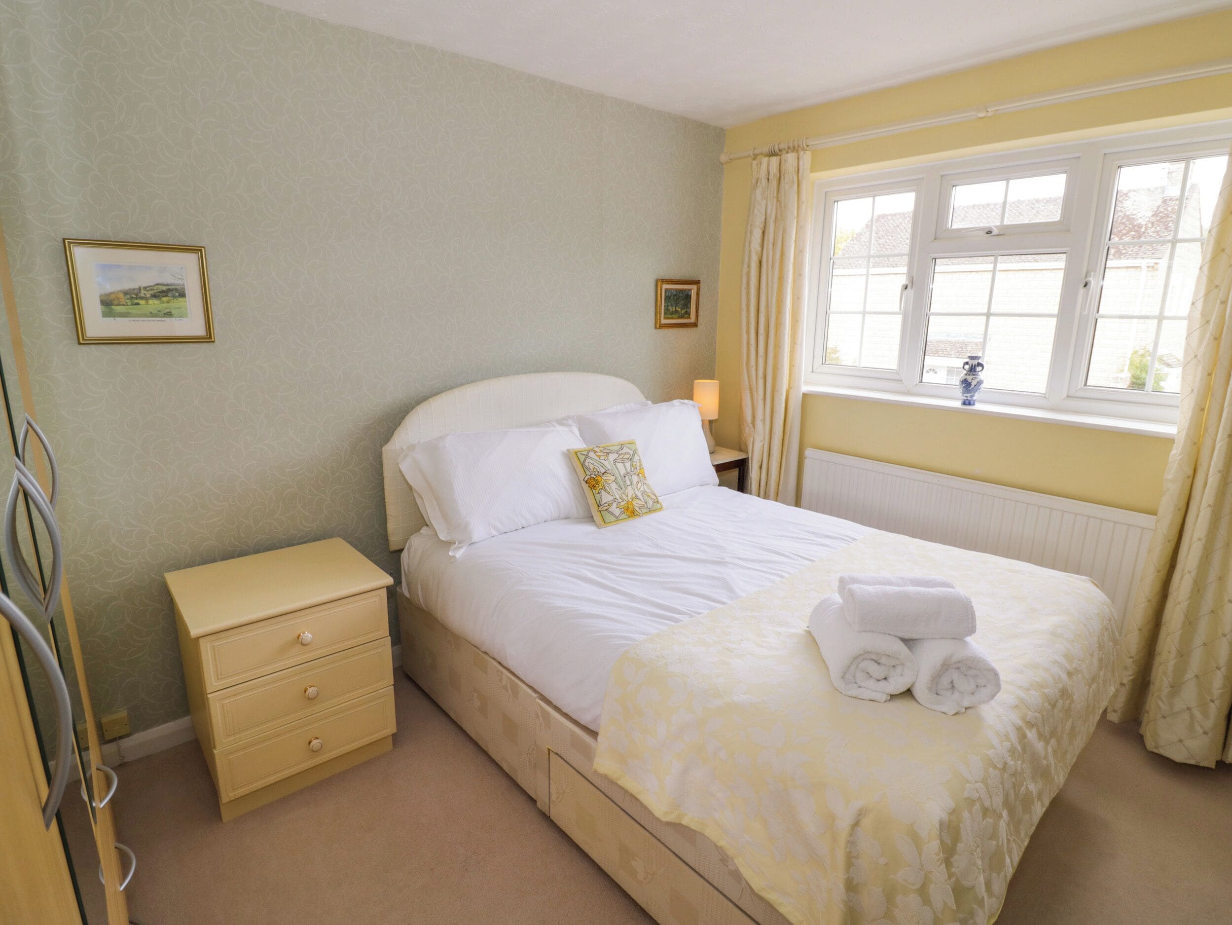 A bedroom with a single bed, a bedside table, a nightstand, a window with curtains, and a radiator.