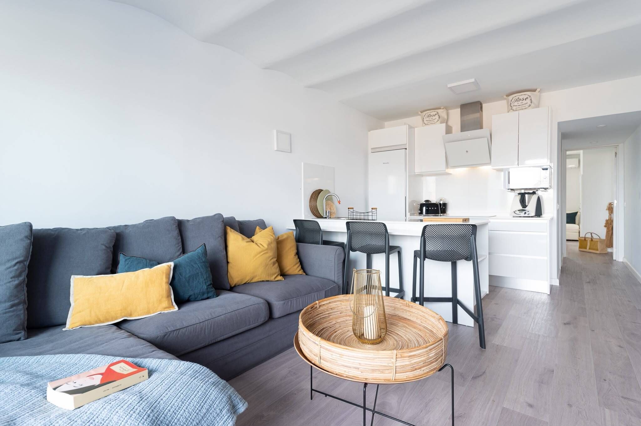 A modern living room with a grey sofa, yellow pillows, a woven coffee table, and a kitchen area with white cabinets and a coffee machine.