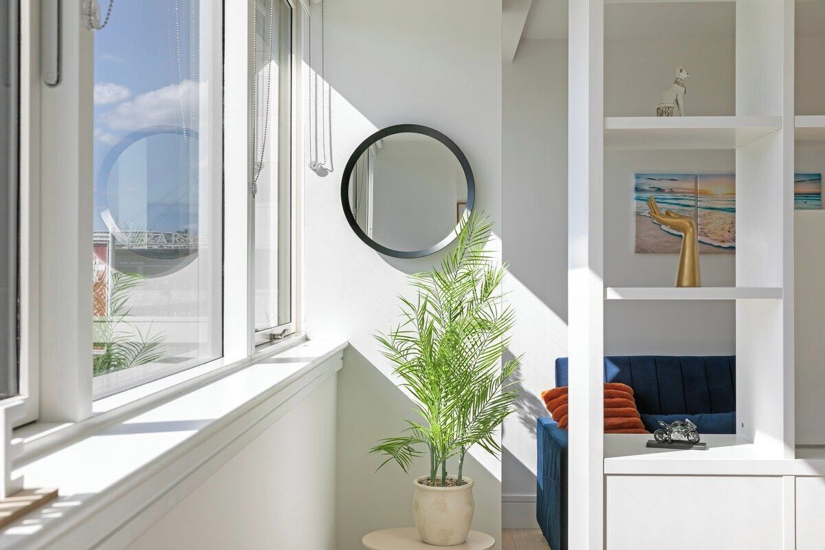 A room with a large window, a potted plant, a mirror, and a blue sofa with orange cushions.