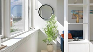 Ein Zimmer mit großem Fenster, Topfpflanze, Spiegel und blauem Sofa mit orangefarbenen Kissen.