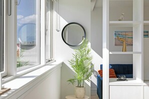 Ett rum med ett stort fönster, en krukväxt, en spegel och en blå soffa med orange kuddar.