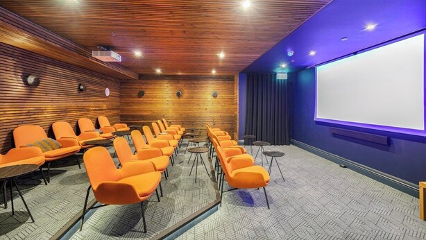 Ein Konferenzraum mit Holzwandverkleidung, orangefarbenen Stühlen, einer Projektionsleinwand und einer blauen Akzentwand.