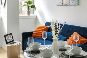 A dining area with a table set for four, a laptop on a side table, and a blue sofa with orange cushions in the background. - Wembley Studio | Cinema, Gym & Workspace (England)