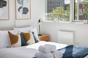 A neatly made bed with white and patterned pillows, a blue blanket, and rolled towels on a window sill. - Wembley Studio | Cinema, Gym & Workspace (England)