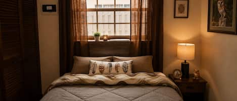 Un dormitorio con cama, ventana con cortinas, lámpara de noche y un cuadro en la pared.
