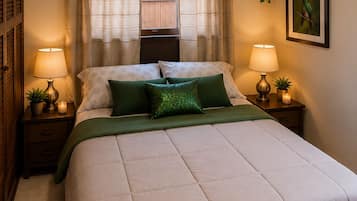 A bedroom with a large bed, two bedside lamps, a window with curtains, an air conditioning unit, and a framed picture on the wall.