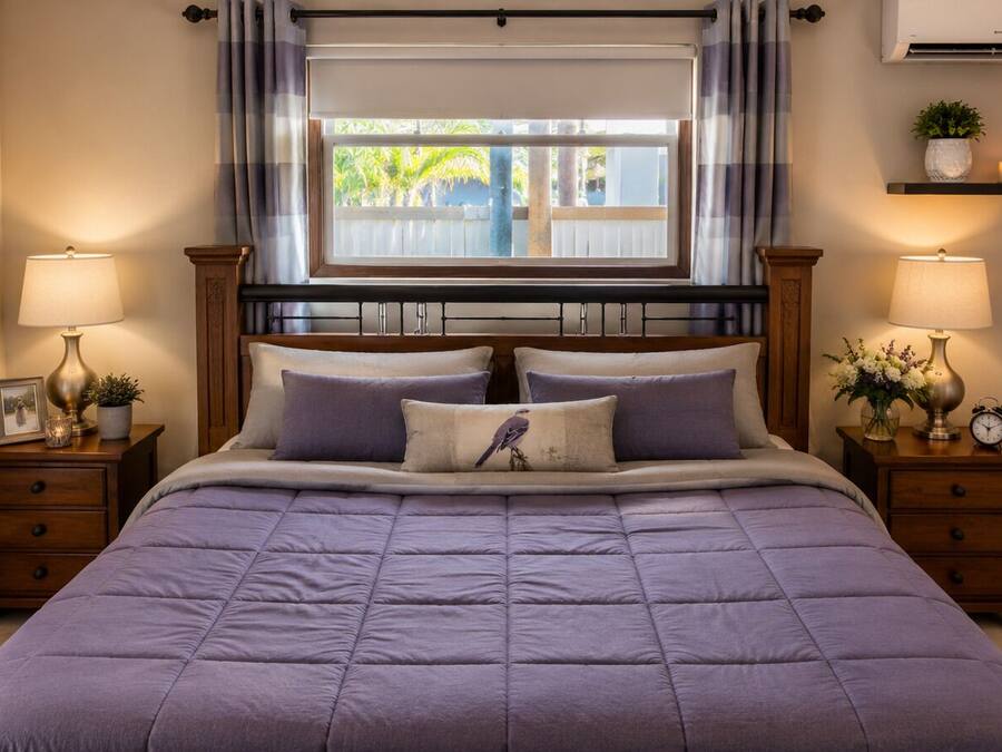 Un dormitorio con una cama, dos mesitas de noche, una mesilla auxiliar, una ventana con cortinas, un aire acondicionado instalado en la pared y un cuadro enmarcado.