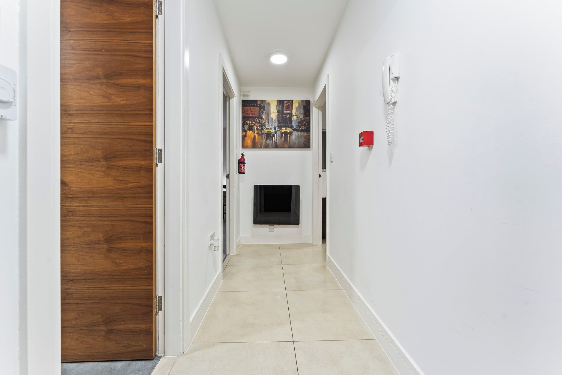 Un couloir avec une porte en bois sur la gauche, un extincteur accroché au mur et un écran de télévision au fond.