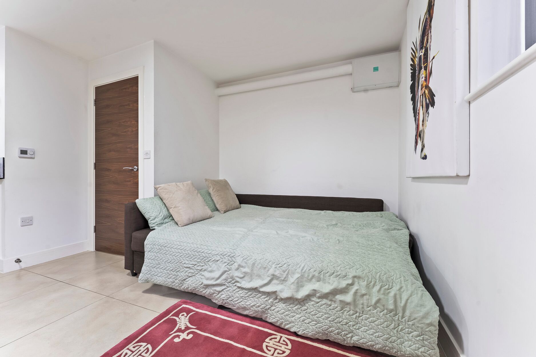 A bedroom with a bed, a door, a wall-mounted air conditioner, and a window with a curtain.
