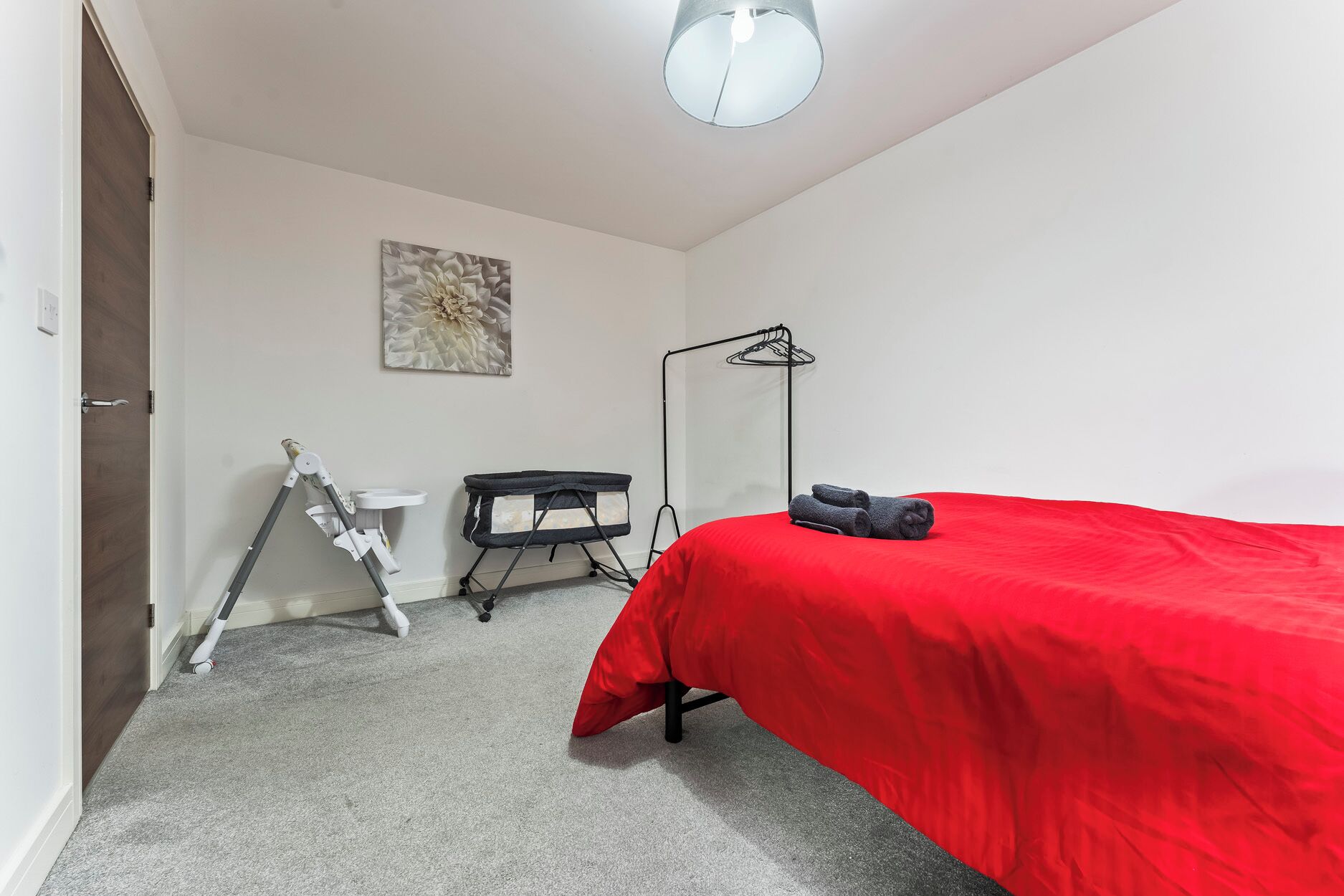 A bedroom with a red bedspread, a white desk, a floor lamp, and a wall-mounted artwork.