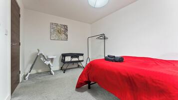 A bedroom with a red bedspread, a white desk, a floor lamp, and a wall-mounted artwork.