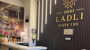 A reception area with a check-in/out sign, a reception desk, a wooden chair, and a wall with vertical slats and a logo. - Shri Ladli Stays Inn (Vrindavan)