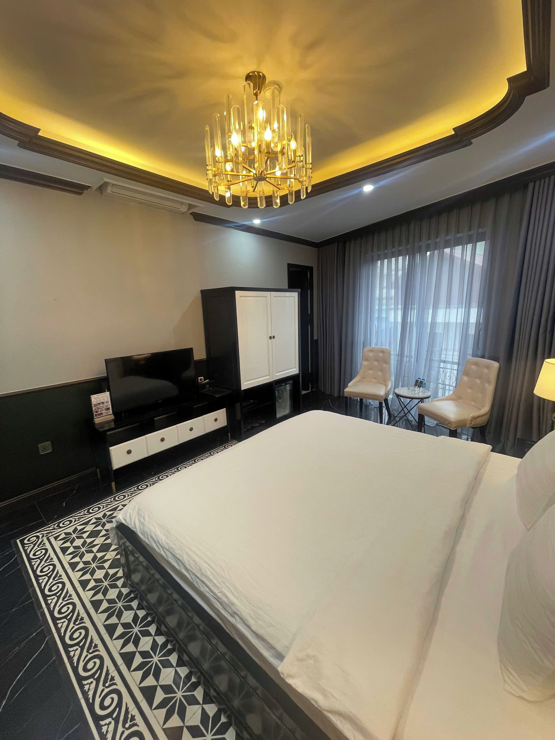A hotel room with a large bed, a TV, a wardrobe, two chairs, and a chandelier.