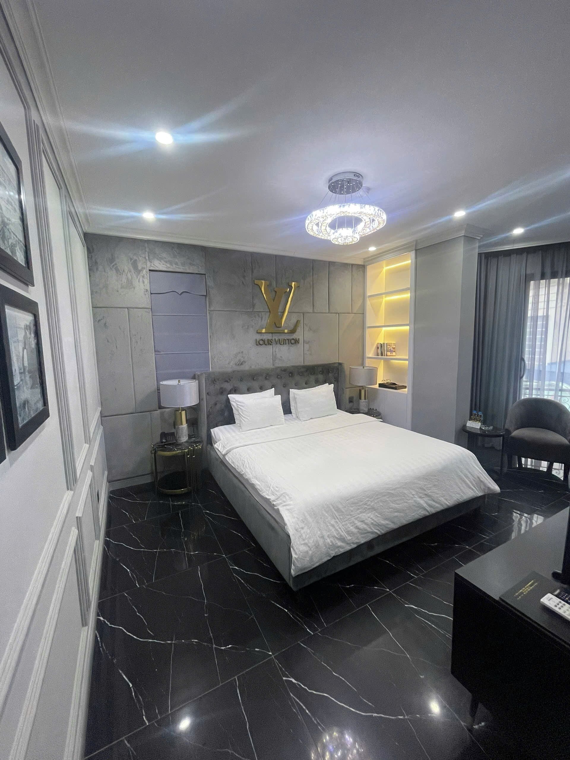 A modern hotel room with a large bed, a Louis Vuitton logo on the wall, a chandelier, and a desk with a lamp.