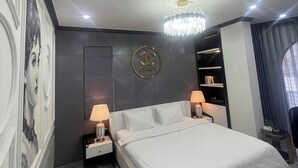 Una habitación de hotel moderna con una cama grande, una araña, un logotipo de Chanel en la pared y una pequeña mesa con lámpara.