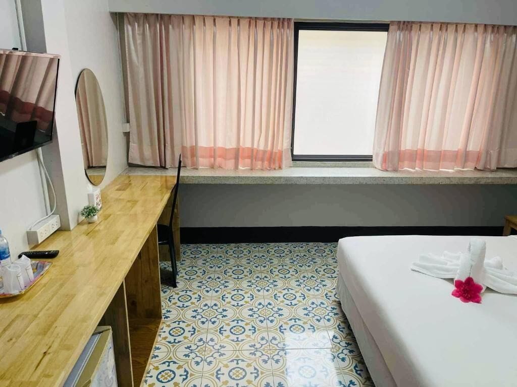 A hotel room with a bed, a desk with a TV, a window with curtains, and a patterned floor.