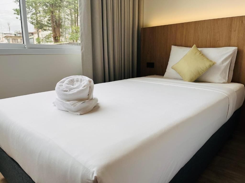 Un lit bien fait, avec une serviette pliée, une tête de lit en bois et une fenêtre avec des rideaux.
