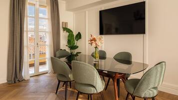 Een eetgedeelte met een glazen tafel, vier stoelen, een vaas met bloemen, een grote flatscreen-tv aan de muur en uitzicht buiten door een glazen deur.
