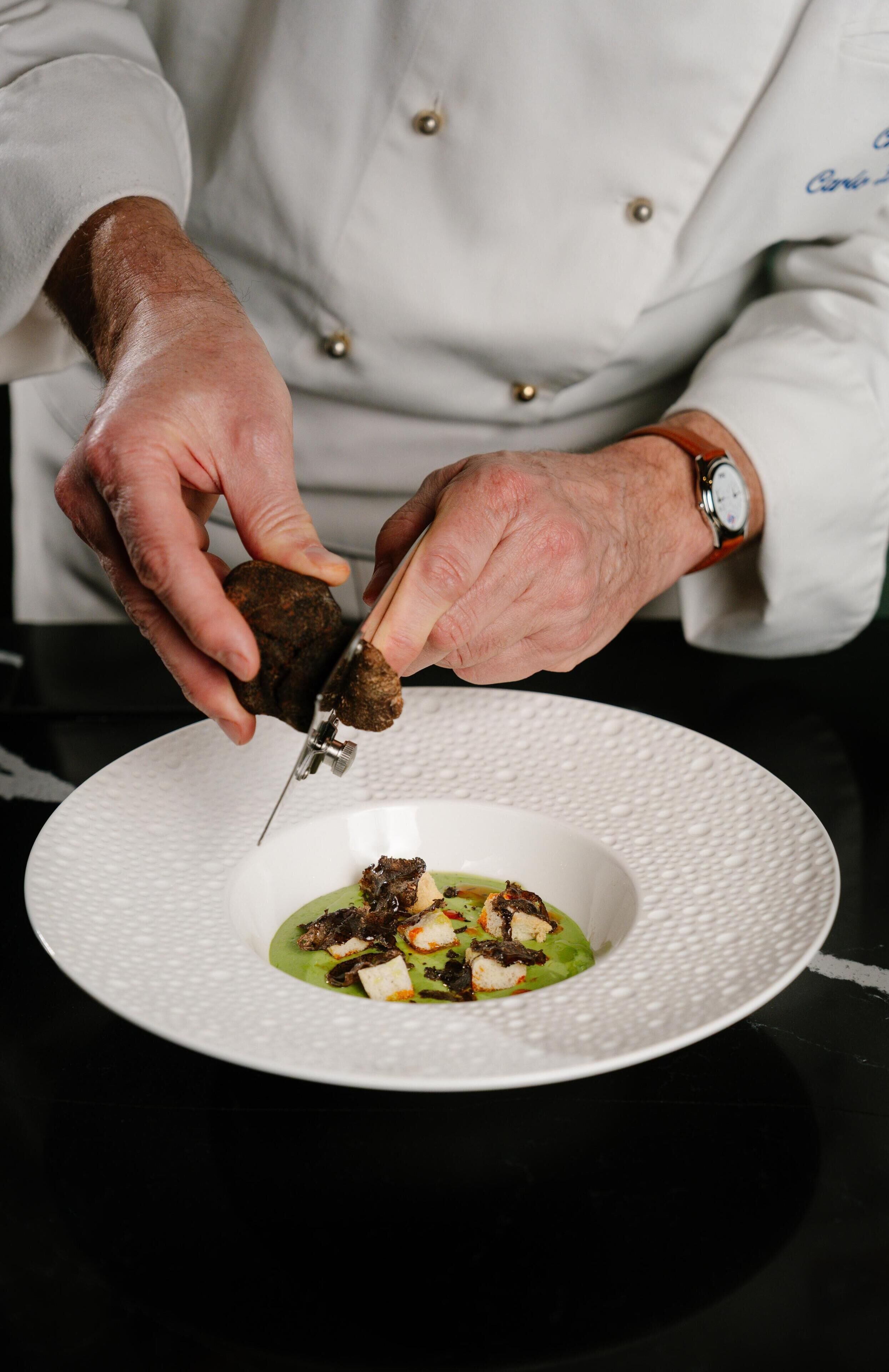 Uma pessoa com avental de chef segurando uma faca, preparando comida em um prato branco com molho verde e croutons.