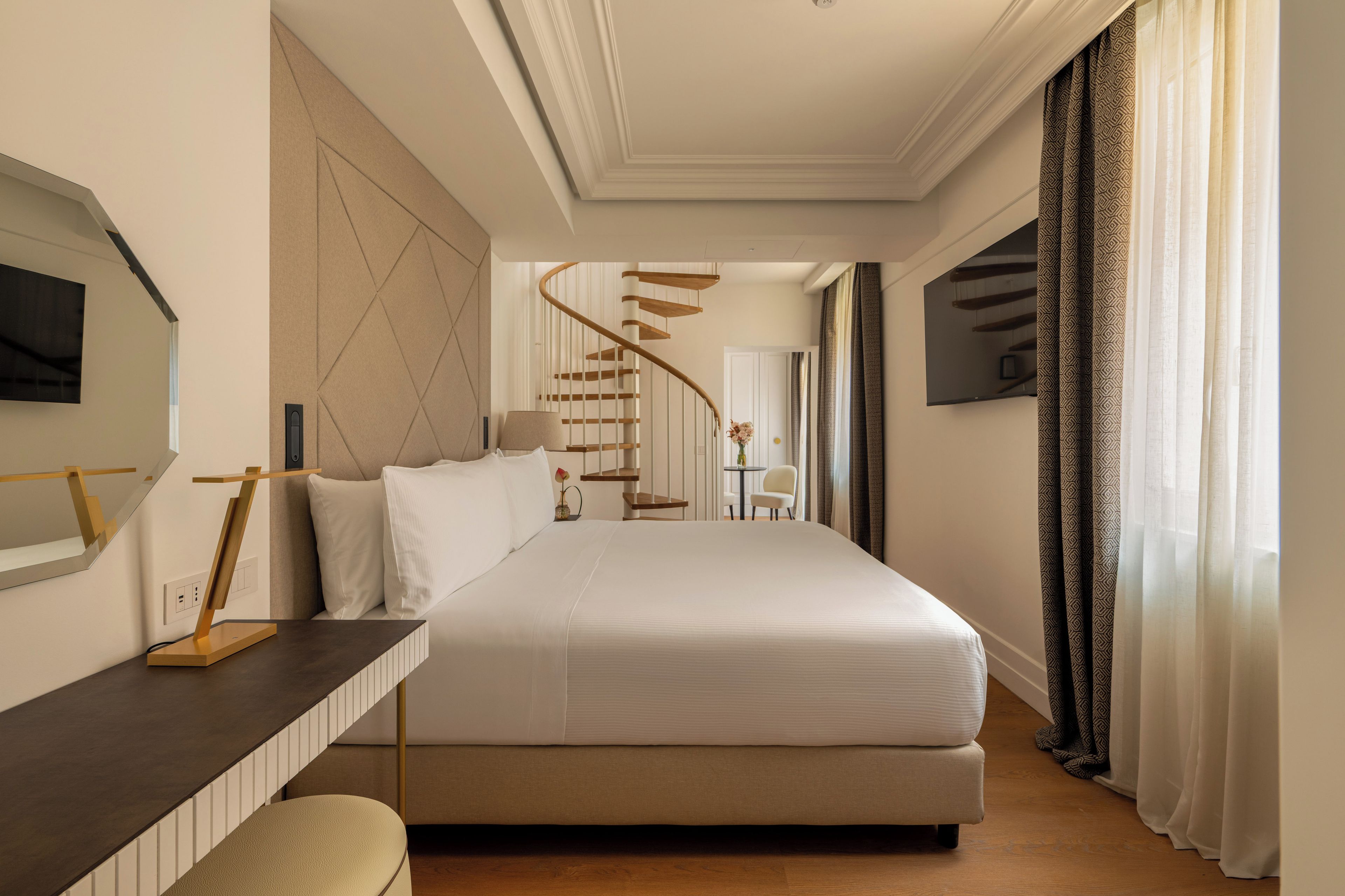 Habitación de hotel con una cama grande, un escritorio con lámpara, un televisor montado en la pared y una escalera de caracol que lleva a otra habitación.