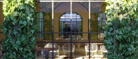 An outdoor seating area with tables and chairs, surrounded by a glass enclosure and covered with ivy.