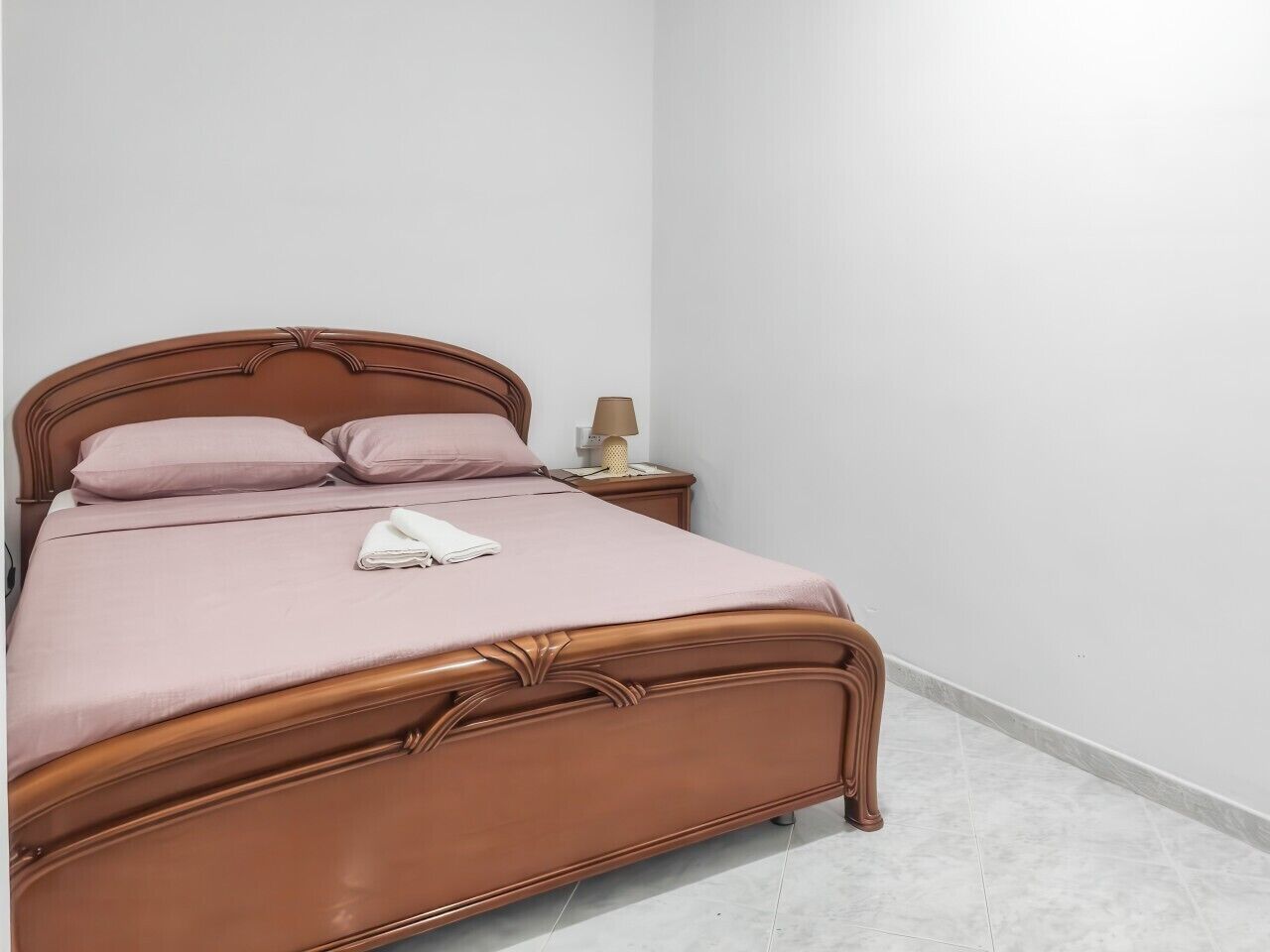 Un lit simple avec un cadre en bois et une couvre-lit rose, une table de chevet avec une lampe, et un mur blanc uni.
