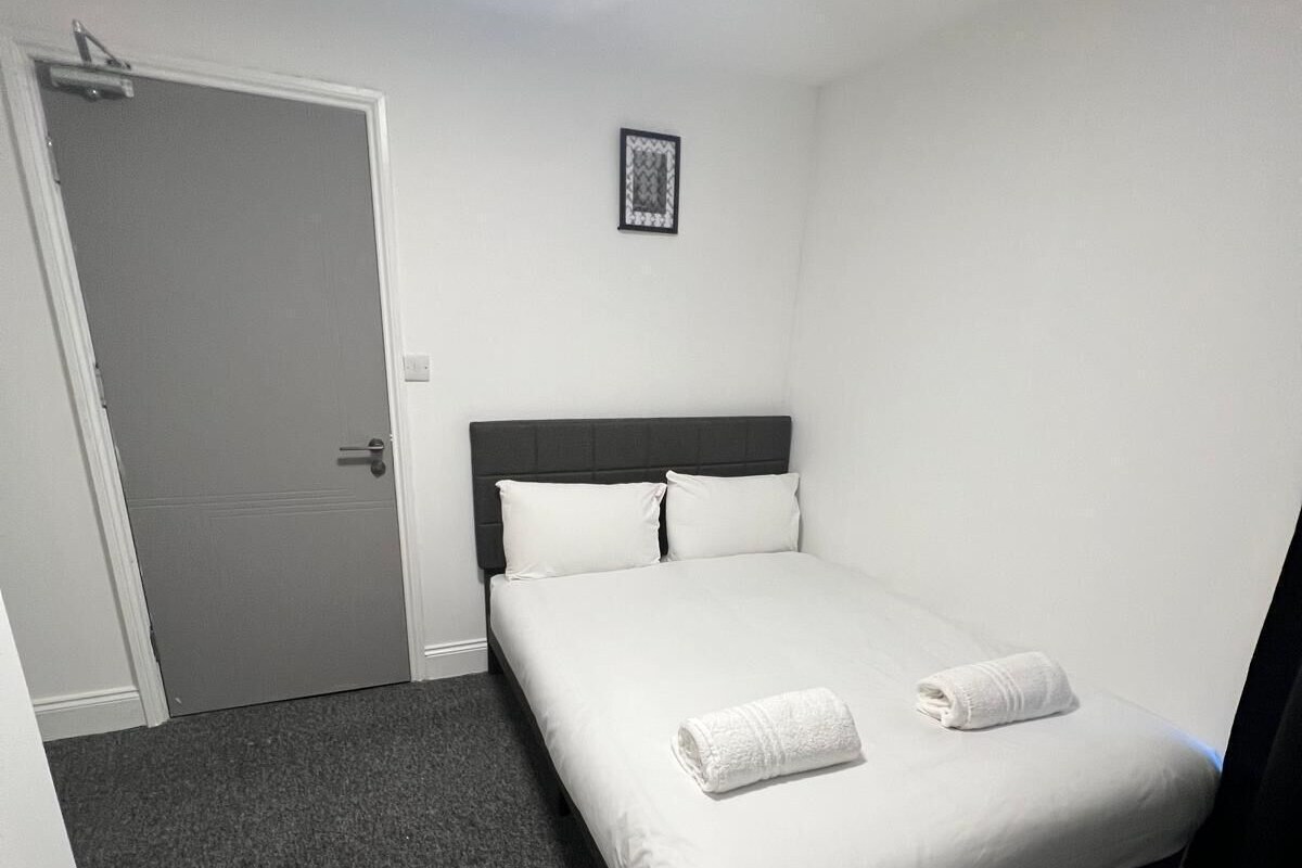 A single bed with white linens and two pillows, a door to the left, and a framed picture on the wall.