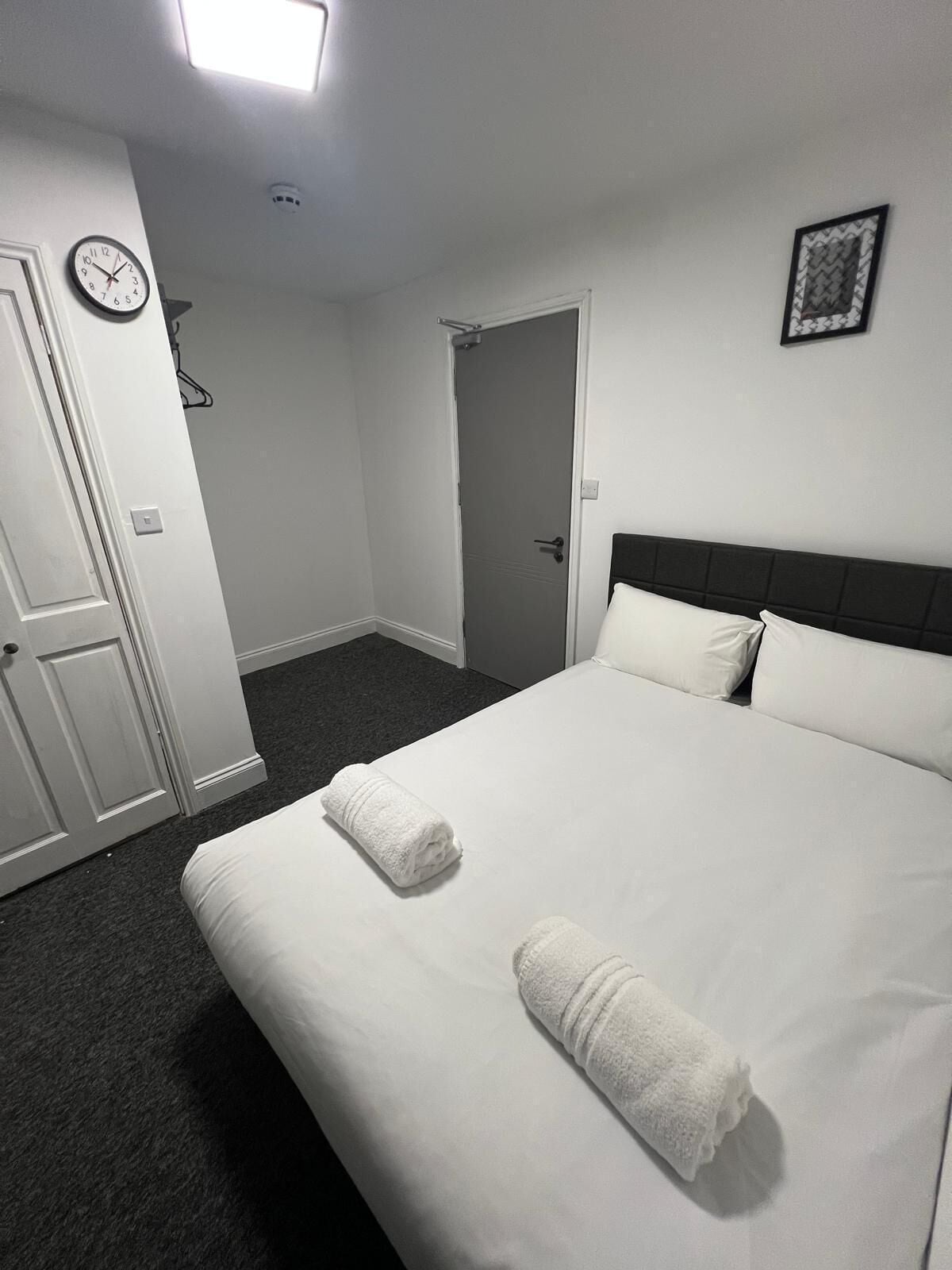 A hotel room with a bed, a door, a clock, and a framed picture on the wall.