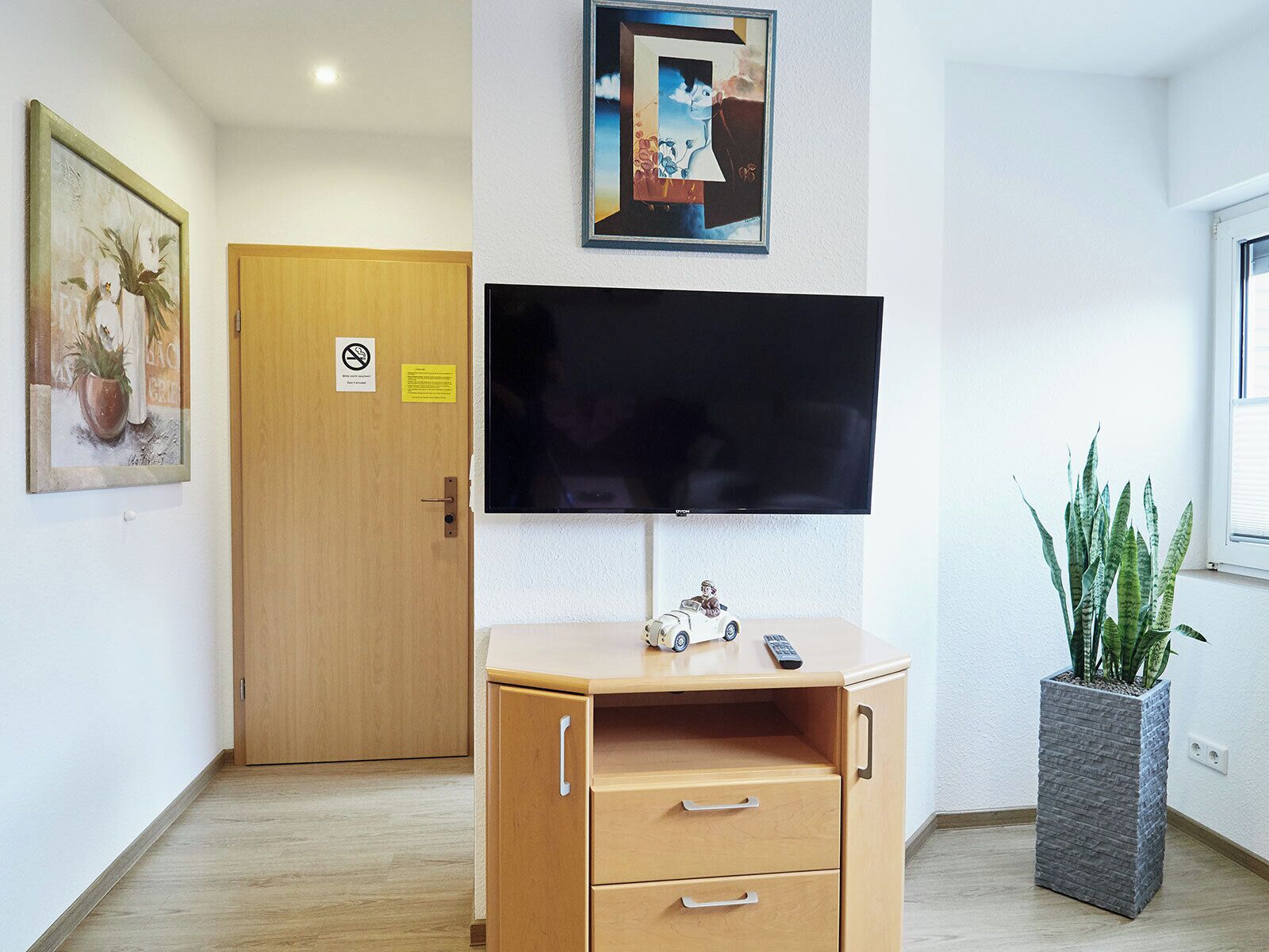 A room with a wooden cabinet, a flat-screen TV mounted on the wall, a door with a no-smoking sign, and a potted plant.